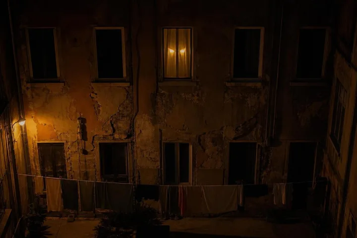 Cortile condominiale buio con una sola finestra accesa e ombre feline sui muri