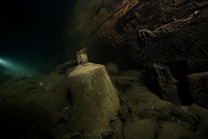 Sedimento scuro sotto una chiglia antica, attraversato da una luce fredda nel buio dell’acqua.