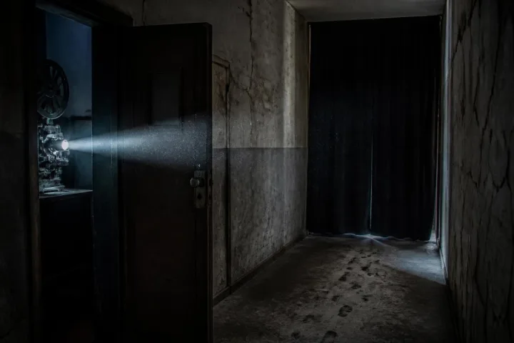 Corridoio tecnico buio dietro una vecchia sala cinematografica con polvere sospesa e una porta di proiezione socchiusa