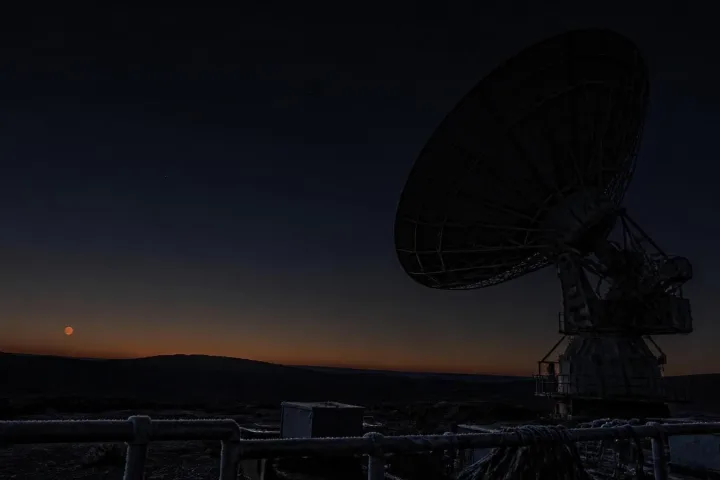Radiotelescopio nel deserto sotto un cielo scuro con Marte basso all'orizzonte