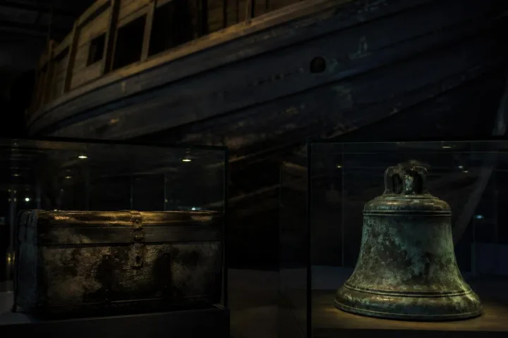 Una campana ossidata e una cassa recuperata da un relitto esposte nella penombra di un museo marittimo.