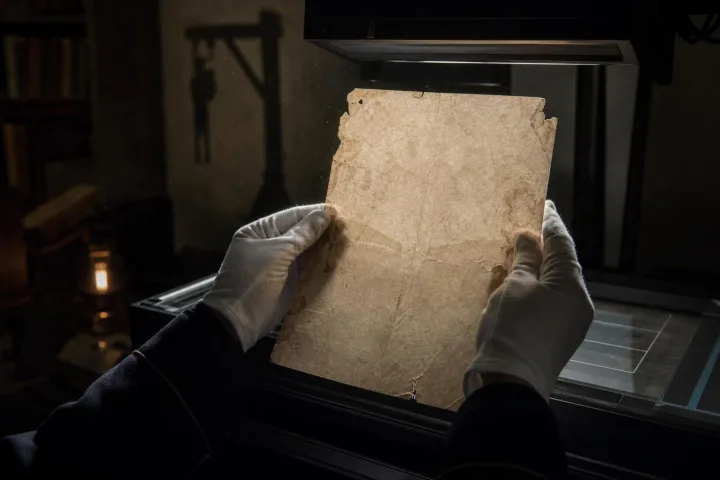 Una pagina d’archivio illuminata sotto uno scanner, con mani guantate e ombre sul muro