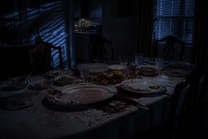 Tavola del Giorno del Ringraziamento abbandonata in una casa buia, con stoviglie rotte e luce blu della polizia dalle persiane