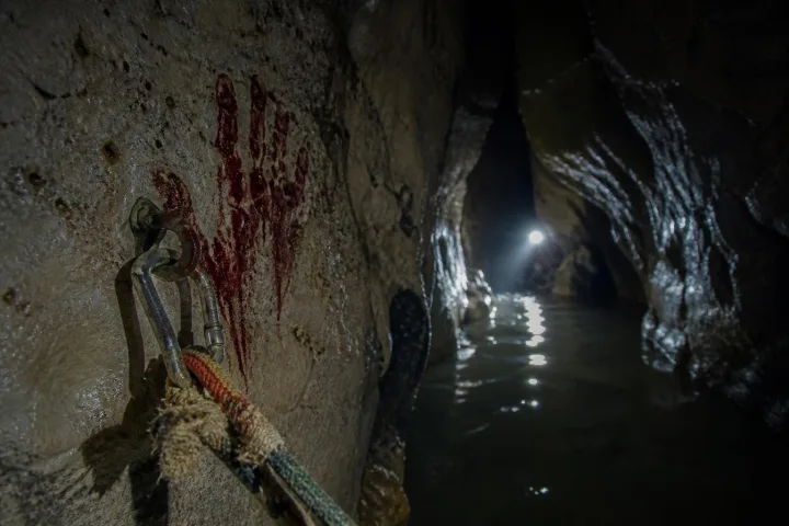 Corda strappata e carabiner nel fango di una grotta allagata, con una luce lontana nel buio