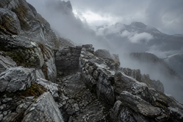 Resti in pietra di una trincea scavata nella roccia di un pendio alpino scosceso, nebbia bassa tra le pietre, luce grigia e fredda del primo mattino.