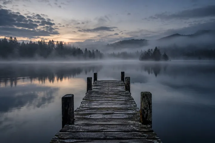 Un pontile di legno consumato si allunga su un'acqua immobile e scura, avvolto da una leggera nebbia all'alba con gli alberi sulla riva opposta.